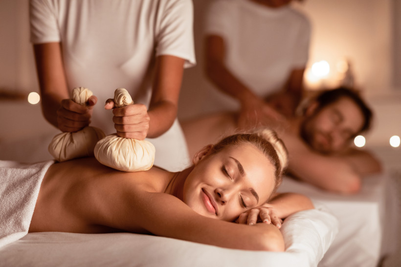 Frau genießt entspannende Massage mit Kräuterstempeln im Spa, im Hintergrund erhält ein Mann eine Rückenmassage