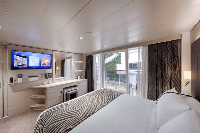 Kabine der MSC World Europa mit Doppelbett, Fernseher und Balkon zur Promenade mit teilweisem Meerblick