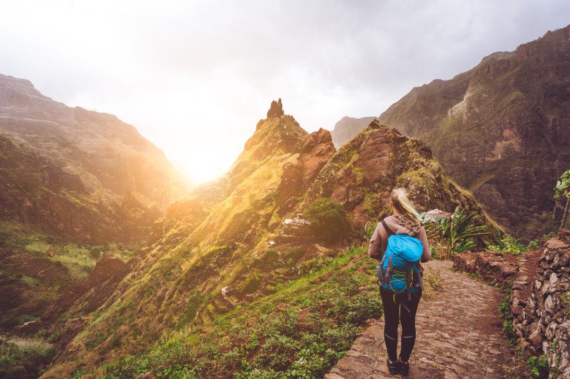 Kapverden Eine Wanderin mit blauem Rucksack steht auf einem schmalen, gepflasterten Bergweg auf den Kapverden und blickt in ein tiefes, grünes Tal. Um sie herum ragen steile, teils bewachsene Felswände in die Höhe. Die Sonne scheint zwischen den Bergen hervor und t