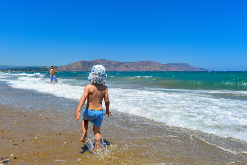 Familienurlaub in Georgioupolis am Strand mit Kind in der Brandung Kind am Strand von Georgioupolis läuft durch seichte Brandung, im Hintergrund Meer und Berge