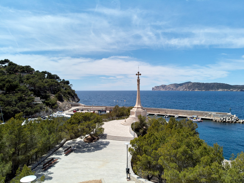 Mallorca Eine weitläufige Promenade mit einem steinernen Denkmal, das eine hohe Säule mit einem Kreuz trägt. Im Hintergrund das tiefblaue Meer mit einem langen Steg, kleinen Booten und einer felsigen Küste. Die Szene ist von grünem Baumbewuchs gesäumt und unter ei