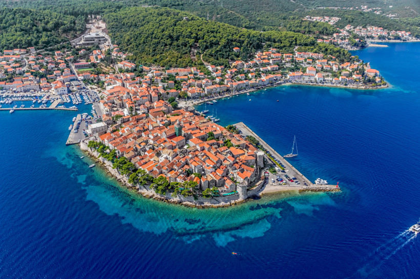 Luftaufnahme der Altstadt von Korčula in Kroatien. Die historische Stadt liegt auf einer Halbinsel und ist von türkisblauem Wasser umgeben. Die roten Ziegeldächer der Häuser und die gut erhaltene Stadtmauer verleihen dem Ort ein mediterranes Flair. Segelb