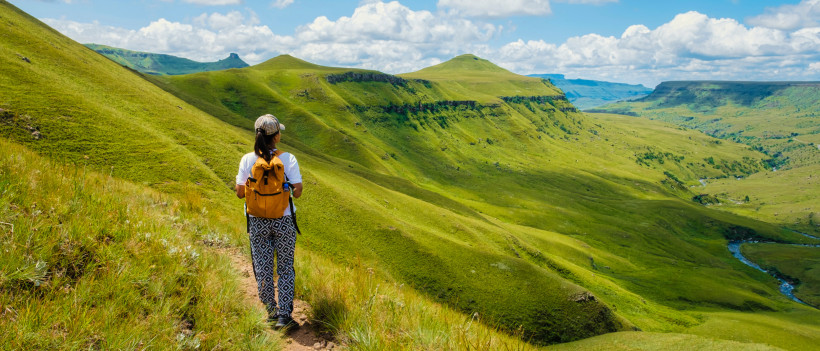 Südafrika Eine Frau mit orangefarbenem Rucksack wandert über einen schmalen Pfad durch grüne Hügel und Berge in Südafrika. Der weite Blick auf die hügelige Landschaft und der blaue Himmel mit weißen Wolken vermitteln Ruhe und Naturerlebnis.