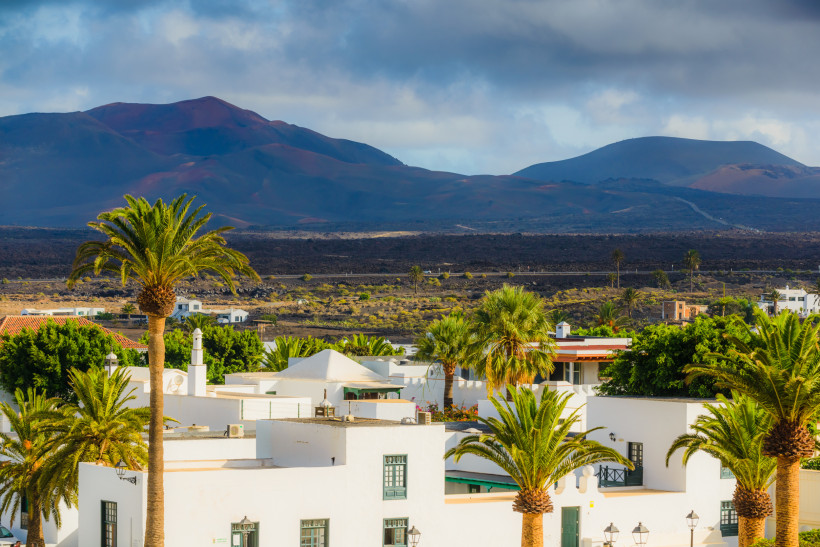 Lanzarote Westen – Yaiza vor der Vulkanlandschaft des Timanfaya Nationalparks Weiß getünchte Häuser und Palmen im Ort Yaiza auf Lanzarote, mit der eindrucksvollen Vulkanlandschaft des Timanfaya Nationalparks im Hintergrund.