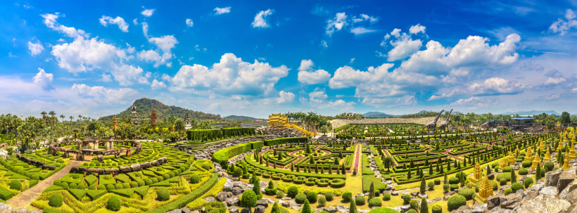 Thailand Panorama eines aufwendig gestalteten botanischen Gartens mit symmetrischen Heckenmustern, tropischen Pflanzen, goldenen Skulpturen und asiatischer Tempelarchitektur. Im Hintergrund bewaldete Hügel und ein blauer Himmel mit wenigen Wolken.