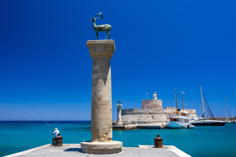 Der Mandraki-Hafen auf Rhodos: Im Vordergrund eine antike Säule mit einer Bronzestatue eines Hirsches, Symbol der Insel. Ein Angler sitzt am Rand des Kais. Im Hintergrund liegt die mittelalterliche Festung Agios Nikolaos, umgeben von Segelbooten und türki