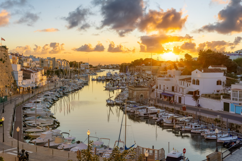 Hafen von Ciutadella de Menorca bei Sonnenuntergang. Zahlreiche Boote und Yachten liegen im Wasser, umgeben von weißen Häusern und alten Stadtmauern.
