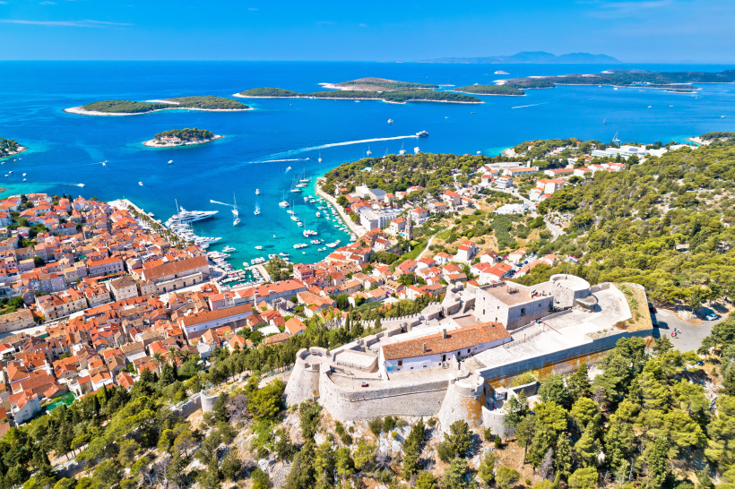 Luftaufnahme von Hvar-Stadt auf der gleichnamigen Insel in Kroatien. Die Altstadt mit ihren roten Ziegeldächern zieht sich entlang der Bucht, in der zahlreiche weiße Boote und Yachten liegen. Im Vordergrund thront die historische Festung Fortica auf einem