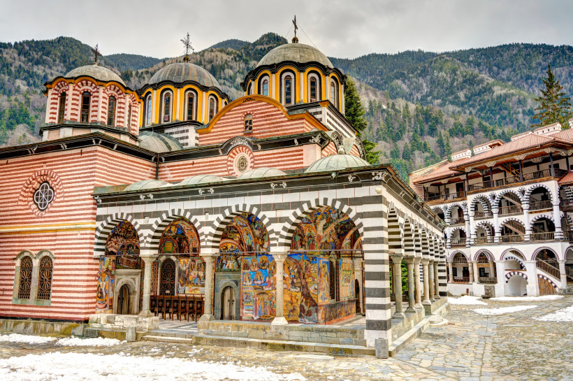 Das Rila-Kloster in Bulgarien mit auffälliger schwarz-weißer Bogenfassade und bunten Wandmalereien. Im Vordergrund die Hauptkirche mit Kuppeln, dahinter umliegende Gebäude mit Holzgalerien. Umgeben von verschneiten Wegen und bewaldeten Bergen.