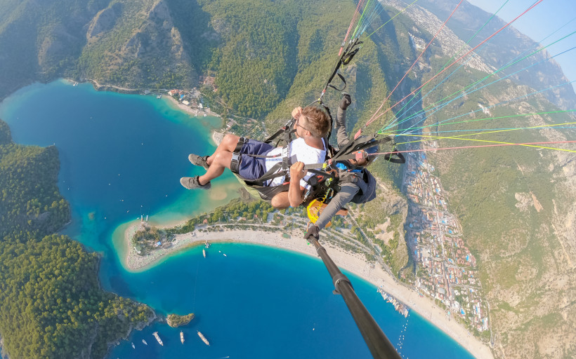 Paragliding über der türkisblauen Lagune von Ölüdeniz in der Türkei. Zwei Personen gleiten im Tandemflug durch die Luft, unter ihnen die grüne Küstenlandschaft, das Meer und kleine Boote.