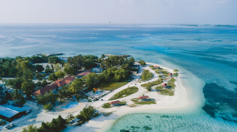 Malediven - Maafushi Drohnenaufnahme einer bewohnten Insel der Malediven mit weißen Sandstränden, Palmen, Häusern und kristallklarem Wasser, das in verschiedenen Blautönen schimmert.