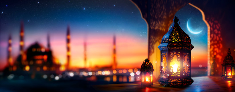 wunderschöne Szene mit traditionellen, orientalischen Laternen, die golden leuchten. Sie stehen auf einem Holzfensterbrett, dessen Muster durch das Licht kunstvolle Schatten werfen. Der Hintergrund ist verschwommen und zeigt eine große Moschee mit mehrere