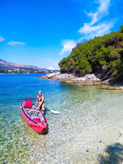 Aktiver Inselgenuss auf Korfu: Eine Frau legt mit ihrem Kajak an einer kleinen, versteckten Bucht mit glasklarem Wasser an. Die felsige Küste ist von dichter Vegetation umgeben, dahinter ziehen sich die Hänge bis in die Ferne. Ein perfekter Ort für indivi