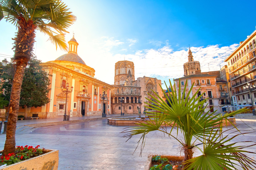 Blick auf einen sonnigen Platz in Valencia mit der barocken Basilika, gotischer Kathedrale und Palmen im Vordergrund. Historische Gebäude umrahmen die Szene.