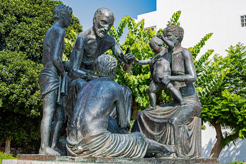 Bronzeskulpturengruppe, die den antiken griechischen Arzt Hippokrates zeigt, umgeben von Zuhörern und einer Frau mit Kind. Die Figuren sitzen und stehen in einer lebendigen Szene, als ob er medizinisches Wissen weitergibt. Im Hintergrund sind grüne Bäume 