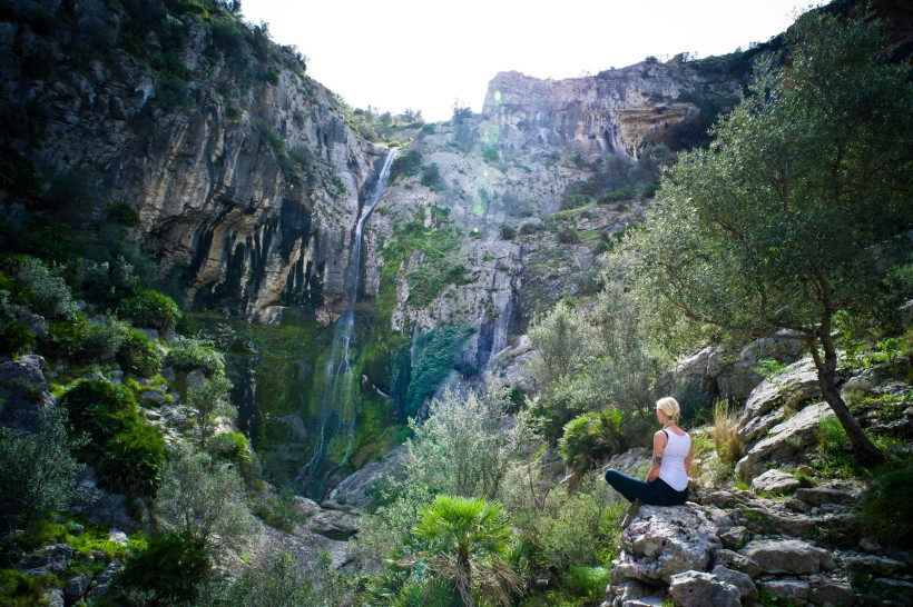 Frau sitzt auf Felsen vor Wasserfall und grüner Berglandschaft beim Wandern auf Teneriffa