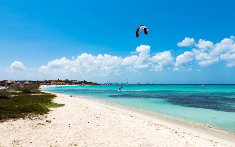 Blick über einen weißen Sandstrand auf Aruba mit türkisblauem Wasser. Ein Kitesurfer gleitet über die Wellen, der Drachen fliegt hoch am blauen Himmel. Im Hintergrund sind weitere Badegäste und Boote zu sehen.