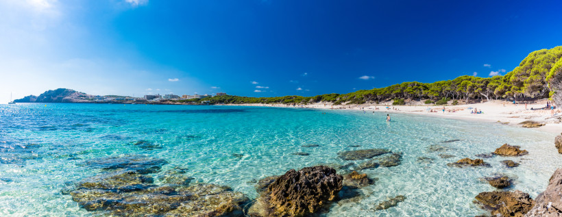 Strand Cala Agulla bei Cala Ratjada auf Mallorca mit kristallklarem, türkisfarbenem Wasser, Felsen im Meer und Pinienwald im Hintergrund – beliebter Naturstrand an der Ostküste