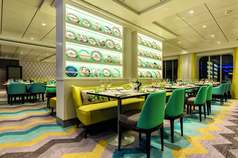Innenansicht eines Restaurants an Bord der Mein Schiff 6 mit gedeckten Tischen, grünen Stühlen und beleuchteten Regalen mit dekorativen Tellern