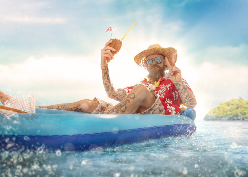 Curaçao Ein tätowierter Mann mit Strohhut, Sonnenbrille und Hawaiihemd liegt entspannt auf einer blauen Luftmatratze im Meer. In der Hand hält er eine Kokosnuss mit Strohhalm und Cocktail-Schirmchen. Er zeigt ein Peace-Zeichen, im Hintergrund scheint die Sonne