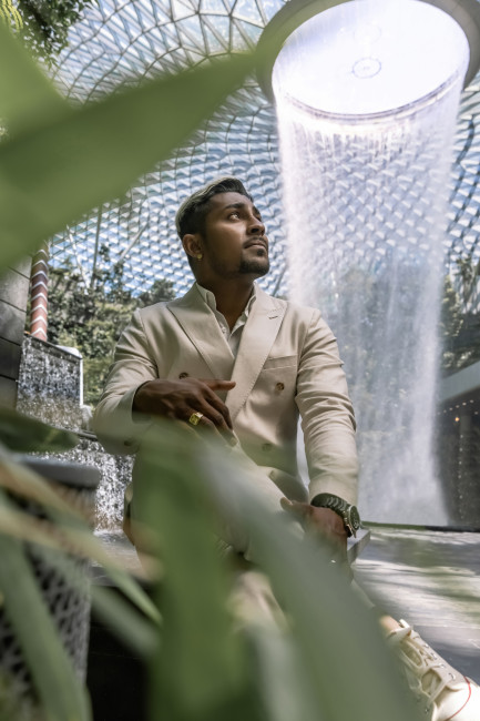 Singapur Ein junger Mann im hellen Anzug sitzt nachdenklich in einer tropischen Umgebung mit üppigem Grün. Hinter ihm fällt ein beeindruckender Indoor-Wasserfall von einer hohen Glaskuppel, durch die natürliches Licht fällt
