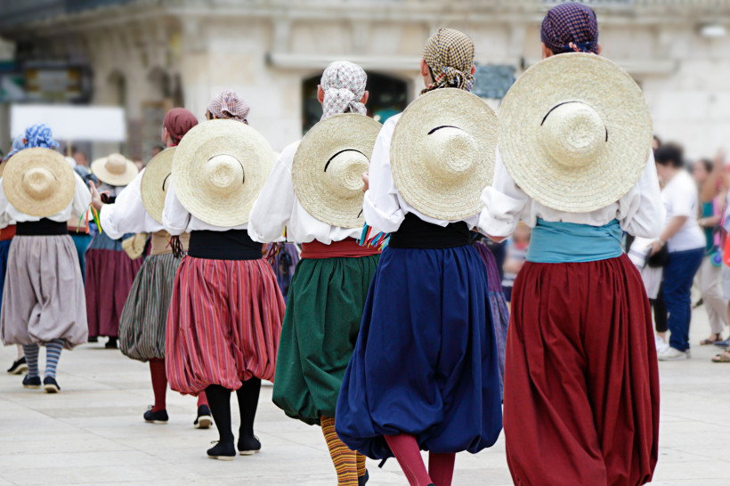 Mallorca Gruppe von Menschen in traditioneller mallorquinischer Tracht, die auf einem öffentlichen Platz tanzen. Alle tragen weite bunte Röcke, weiße Blusen und Strohhüte auf dem Rücken. Die Szene spielt sich im Freien ab, umgeben von anderen Passanten.