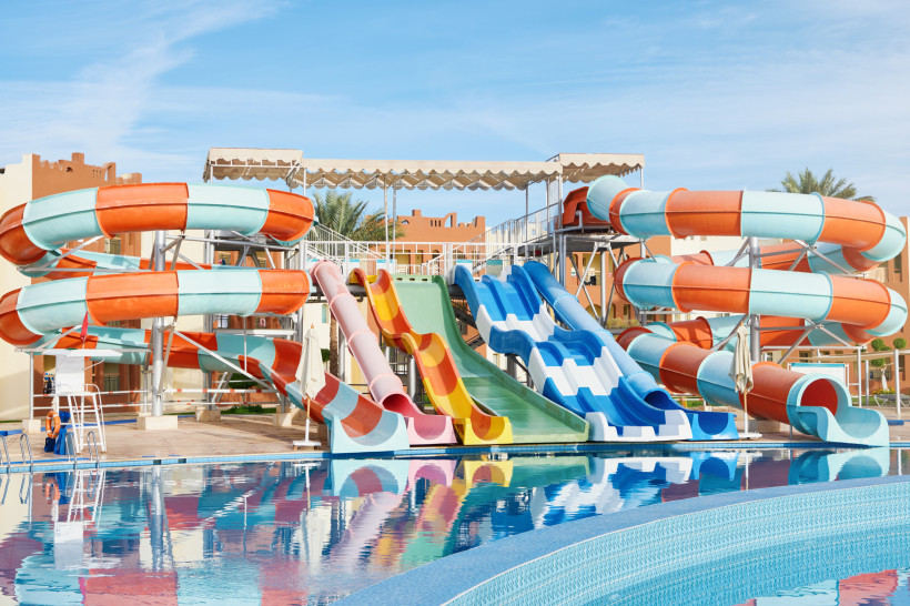 Ägypten - Marsa Alam Großer Wasserpark mit bunten Wasserrutschen, die in einen klaren Pool führen. Im Hintergrund stehen Hotelgebäude, der Himmel ist blau und wolkenlos.