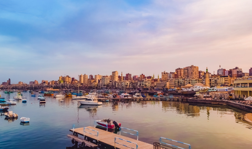Das Bild zeigt den Hafen von Alexandria in Ägypten in der Abenddämmerung. Im ruhigen Wasser liegen zahlreiche bunte Boote, kleinere Yachten und Fischerboote vor Anker. Die Stadt erstreckt sich im Hintergrund mit einer dichten Skyline aus mehrstöckigen Woh
