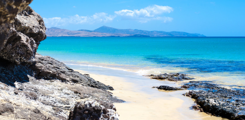 Blick von einem Sandstrand mit Felsen auf den Atlantik und eine Bergkette am Horizont