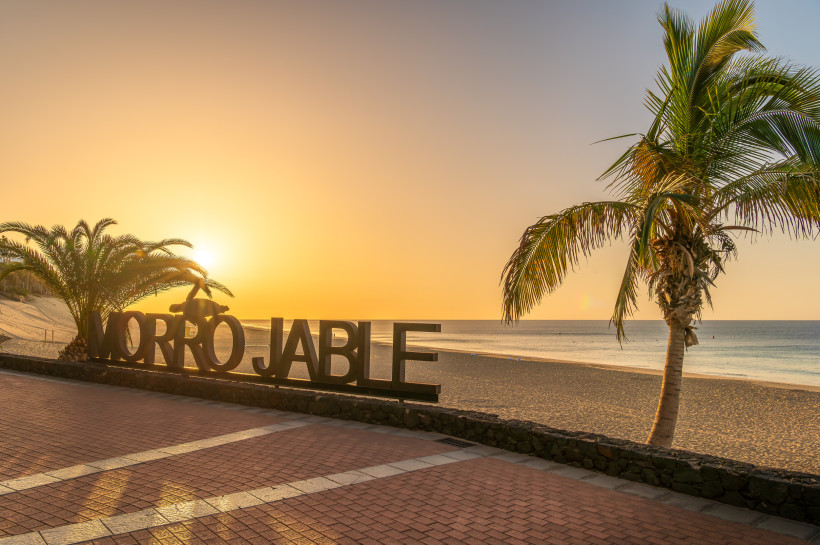 Schriftzug „Morro Jable“ an der Strandpromenade bei Sonnenuntergang, Palmen und Meer im Hintergrund