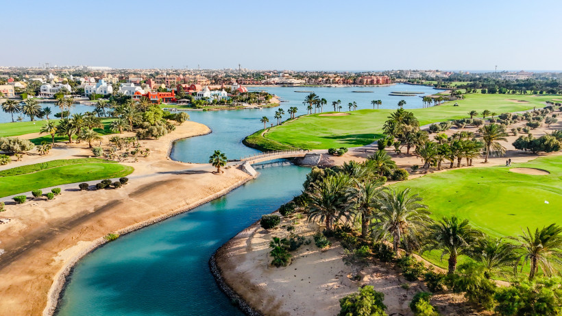 Ägypten - El Gouna Blick auf eine gepflegte Golfanlage in El Gouna, Ägypten. Zwischen sattem Grün und Palmen verlaufen künstliche Wasserkanäle. Im Hintergrund sind bunte Villen und Häuser entlang der Lagunen zu sehen, mit der Stadt El Gouna in weiter Ferne unter blauem Himm