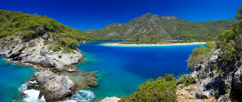 Türkei Diese spektakuläre Küstenaufnahme zeigt eine ruhige Bucht mit intensiv blauem Wasser, eingerahmt von grünen Pinienwäldern und markanten Felsformationen. Der helle Sandstrand in der Ferne bietet sonnige Ruheoasen, während das klare Meer zu Badeabenteuern e