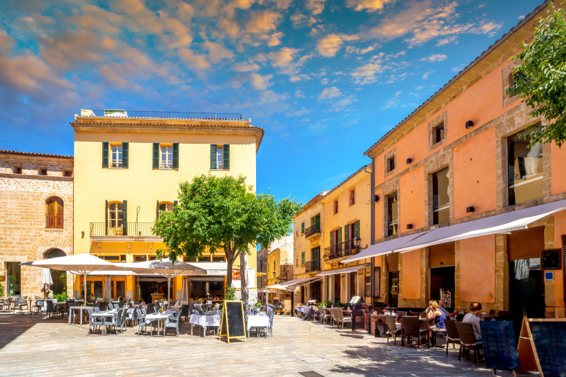 Zentraler Platz in der Altstadt von Alcudia mit Natursteinpflaster, historischen Fassaden und Außenterrassen von Cafés zwischen den Stadtmauern