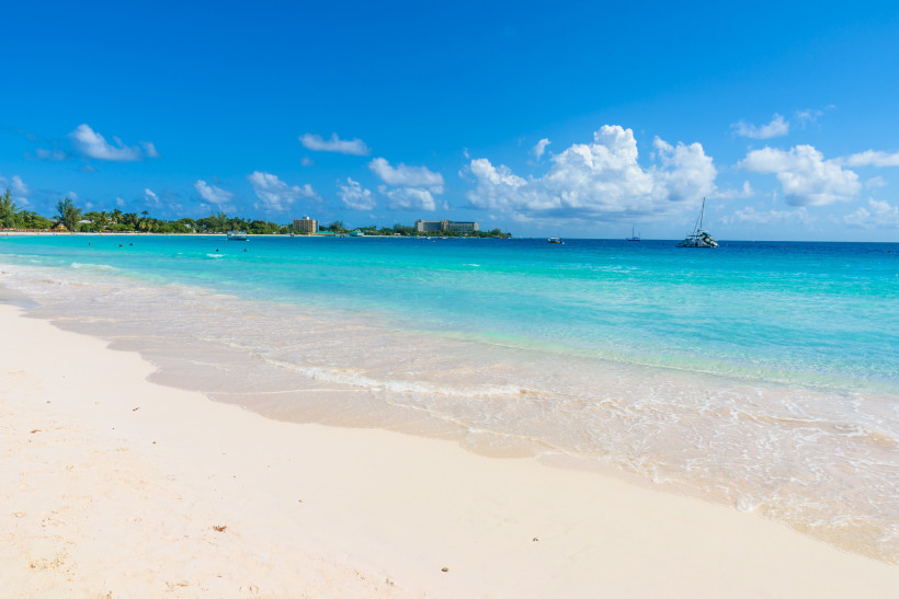 Weißer Sandstrand mit türkisblauem Meer, Palmen und Booten im Hintergrund auf Barbados.