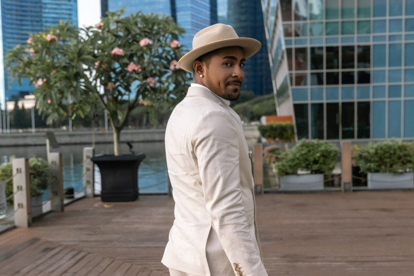 Singapur Ein stilvoll gekleideter Mann in einem beigen Anzug und Hut blickt lächelnd über die Schulter, während er auf einem Holzsteg in einer modernen Stadtlandschaft steht. Im Hintergrund sind ein blühender Baum, moderne Hochhäuser mit Glasfassaden und ein ruhig