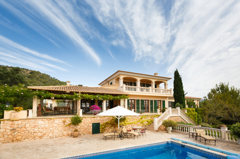 Mallorca Große mediterrane Villa mit mehreren Stockwerken, großzügigem Balkon und überdachter Terrasse. Vor dem Haus befindet sich ein Pool mit Liegestühlen und Sonnenschirm, umgeben von Steinmauern und Zierpflanzen. Der Himmel ist blau mit leichten Schleierwolken