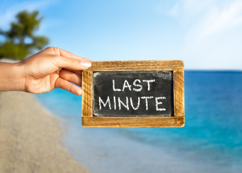 Hand hält eine kleine Tafel mit der Aufschrift „Last Minute“ vor einem Strand und dem türkisblauen Meer.