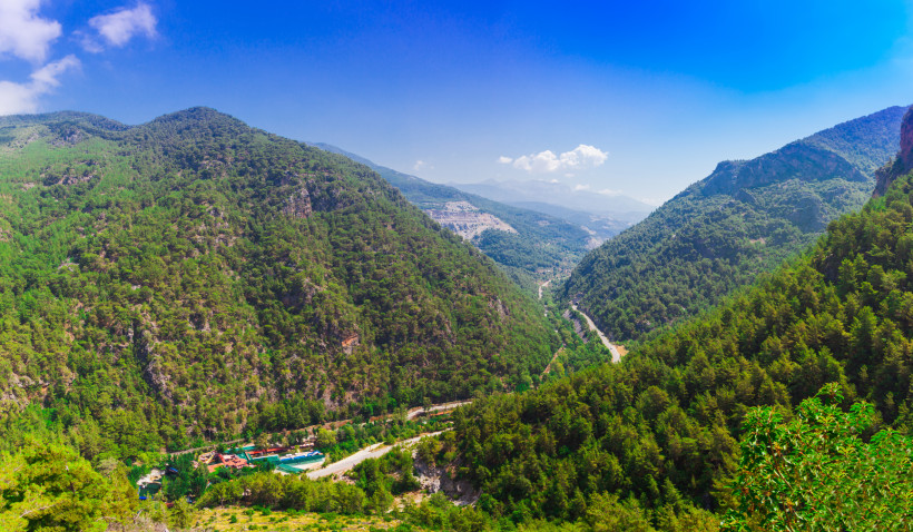 Weitwinkelaufnahme eines bewaldeten Gebirgstals bei Alanya mit kurviger Straße, umliegenden Hügeln und Blick auf das Taurusgebirge im Hintergrund.