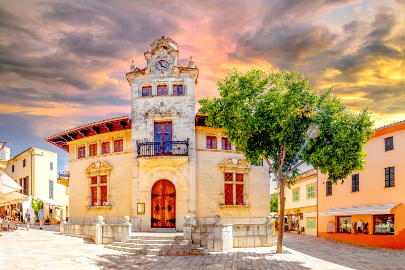 Mallorca - Alcúdia Altstadt Blick auf das historische Rathaus Casa Consistorial in Alcúdia Altstadt auf Mallorca. Das Gebäude ist aus hellem Naturstein erbaut und hat eine dekorative Fassade mit geschnitzten Balkonen, Fensterläden und einem markanten Uhrturm mit Zinnen. Davor führt