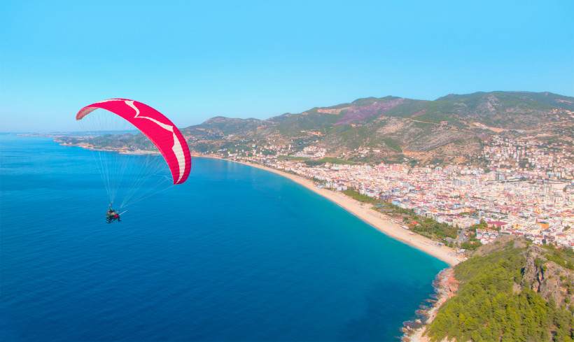 Zwei Personen beim Paragliding mit rotem Gleitschirm über der Küste von Alanya, mit Blick auf das blaue Mittelmeer und die Stadtlandschaft