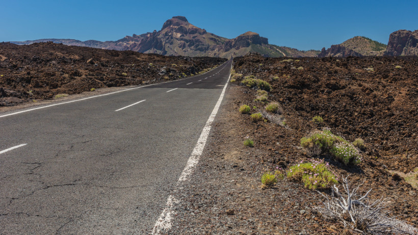Straßenabenteuer durch die Vulkanlandschaft von Teneriffa – ideal für Roadtrips Gerade Straße durch eine karge Vulkanlandschaft mit Blick auf die Berge auf Teneriffa, unter klarem blauen Himmel