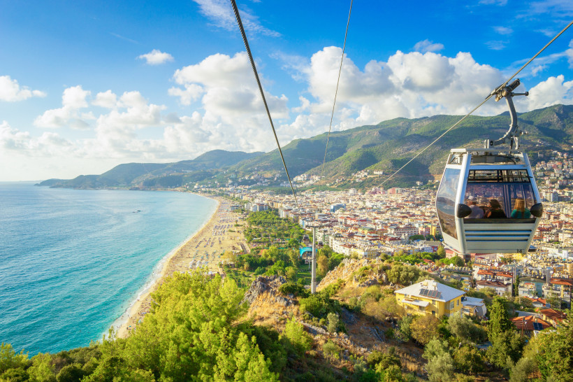 Panoramablick auf Alanya und den Kleopatra-Strand aus einer Seilbahnkabine, die über die Stadt und Küstenlinie führt, mit Bergen im Hintergrund.