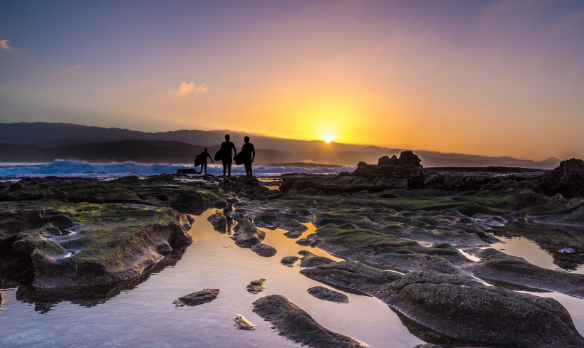 Sonnenuntergang an der Küste von El Confital mit Felsen, Gezeitentümpeln und Silhouetten von drei Surfern