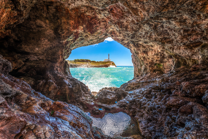 Blick aus einer Felsenhöhle auf das Meer und den Leuchtturm Far de Portocolom auf einer Landzunge