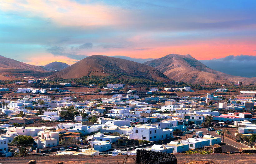 Blick auf das Dorf Yaiza auf Lanzarote mit weißen Häusern, Vulkanlandschaft und Bergen im Hintergrund bei Sonnenuntergang.