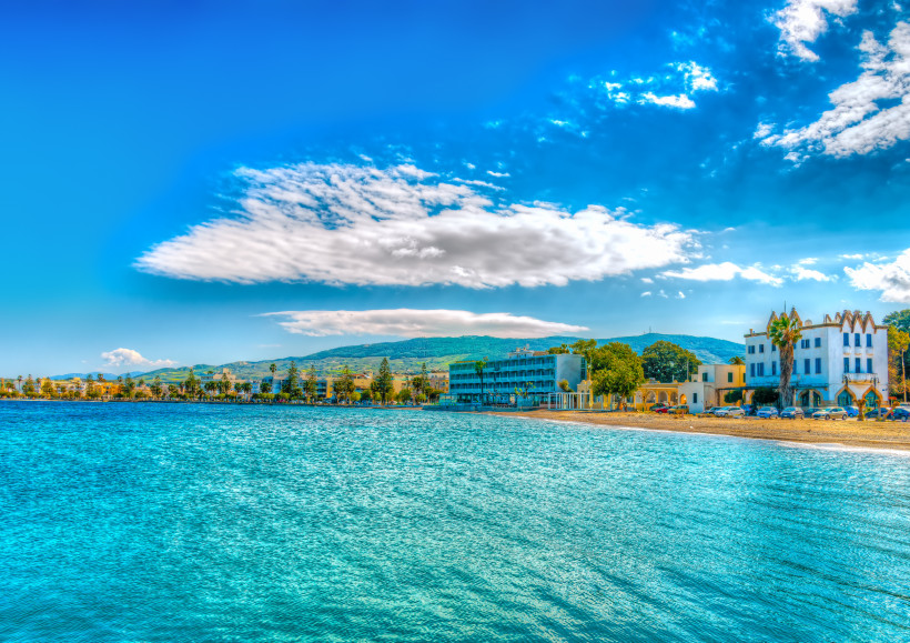 Kos Blick auf die Uferpromenade von Kos-Stadt bei Sonnenschein. Im Vordergrund glitzert das türkisfarbene Wasser der Ägäis. Am Ufer stehen mehrere Gebäude, darunter ein modernes Hotel mit Balkonen und ein weißes, historisch wirkendes Gebäude mit Palmen davor.