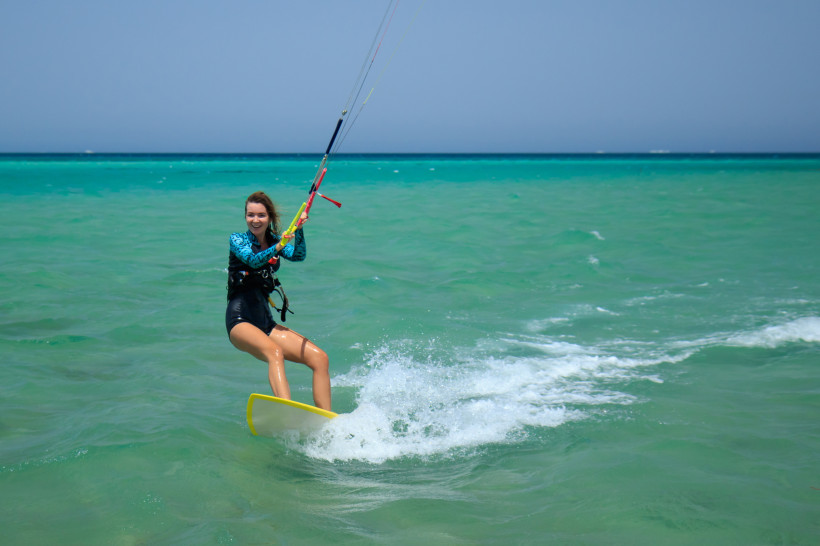 Ägypten - Makadi Bay Eine junge Frau fährt lachend auf einem gelben Kiteboard über das türkisfarbene Meer. Sie hält sich an den Lenkstangen des Kites fest, während Wasser hinter ihr aufspritzt. Der Horizont ist klar, der Himmel wolkenlos – ein perfekter Moment voller Action,