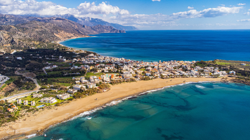 Luftaufnahme eines Küstenortes auf Kreta mit Sandstrand, blauem Meer und weißen Häusern entlang der Küste