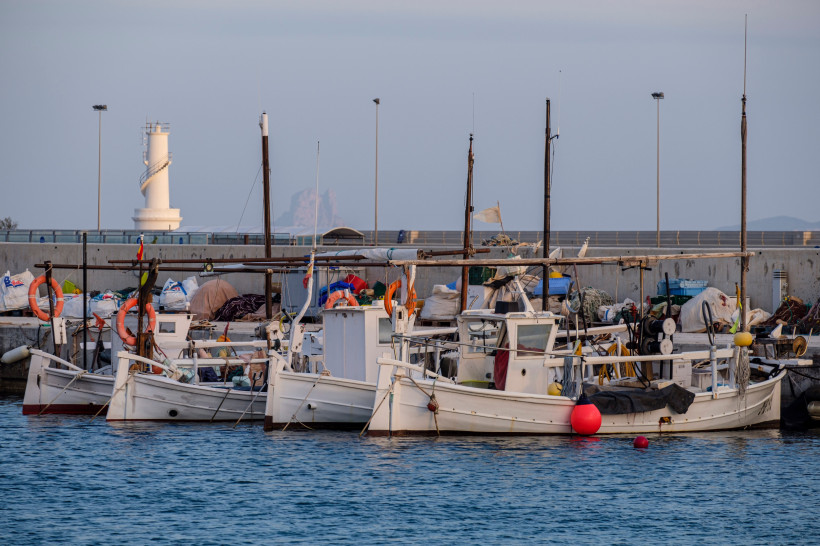 Formentera Mehrere kleine weiße Fischerboote im Hafen, ausgestattet mit Netzen, Bojen und Ausrüstung. Im Hintergrund steht derselbe weiße Leuchtturm, dahinter eine Hafenmauer und ein Felsen im Meer