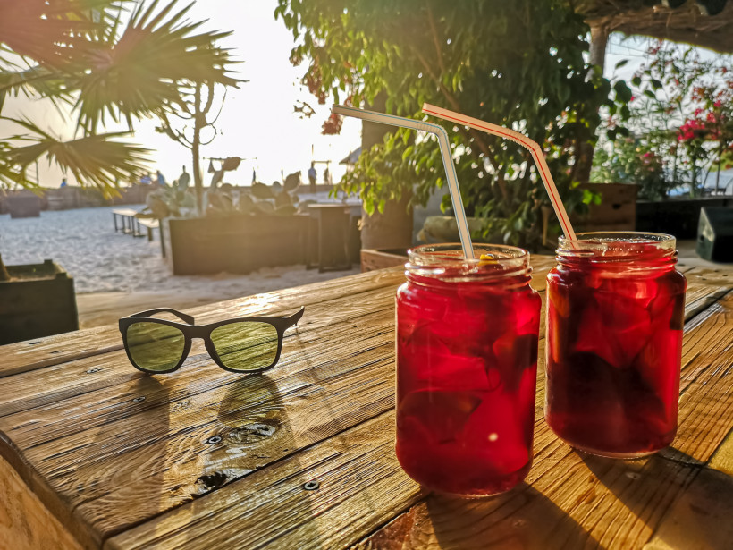 Zwei Gläser mit eisgekühltem roten Getränk und Trinkhalmen stehen auf einem Holztisch in einer Strandbar bei Sonnenuntergang. Daneben liegt eine Sonnenbrille, im Hintergrund sind Strand, Palmen und Menschen im Gegenlicht zu sehen.